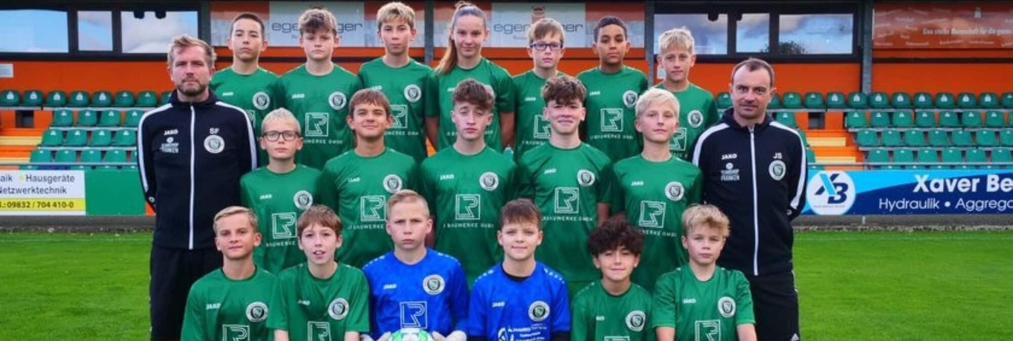 Eine Fußballmannschaft posiert auf einem Rasenplatz. Sie trägt grüne Trikots, drei Spieler tragen rote Torhüter-Outfits. Dahinter ist eine Reihe leerer Stadionsitze und ein Hintergrund mit verschiedenen Werbespots. Der Himmel ist klar und sonnig.