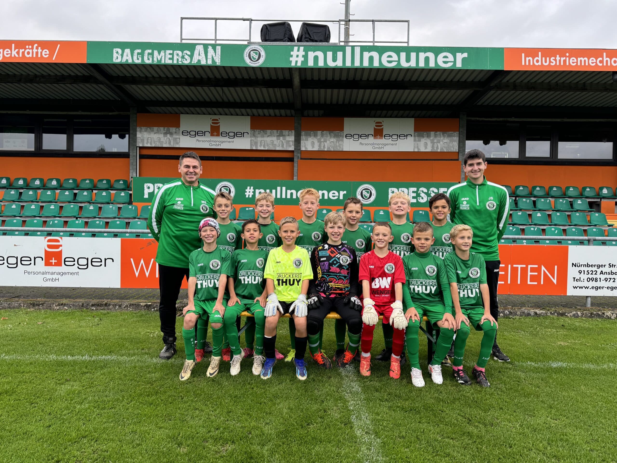 Eine Jugendfußballmannschaft posiert für ein Gruppenfoto auf einem Fußballfeld. Die Mannschaft, in grün-roten Trikots gekleidet, sitzt und steht mit ihren Trainern in zwei Reihen. Dahinter befindet sich ein kleines Stadion mit grünen Sitzen und Werbebannern.