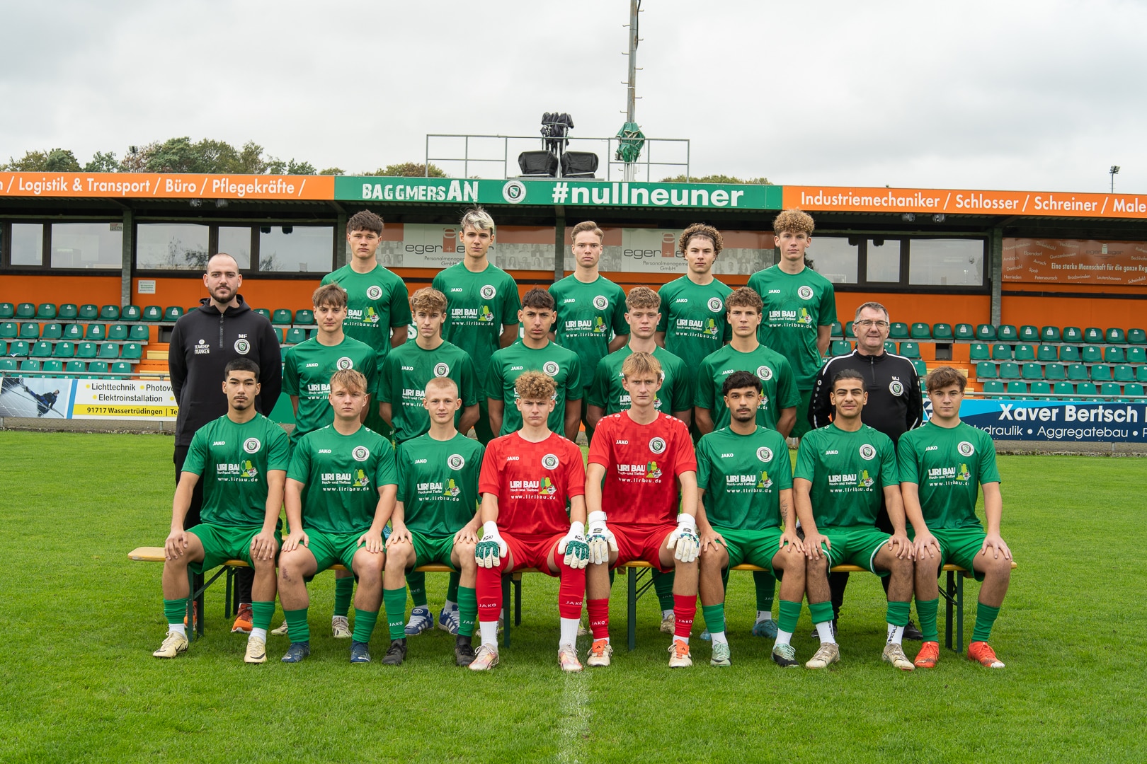 Ein Gruppenfoto einer Jugendfußballmannschaft in grünen Uniformen. Sie posieren in drei Reihen auf einem Feld mit leeren Stadionsitzen im Hintergrund. Das Team besteht aus einer Mischung junger Spieler und Trainer.