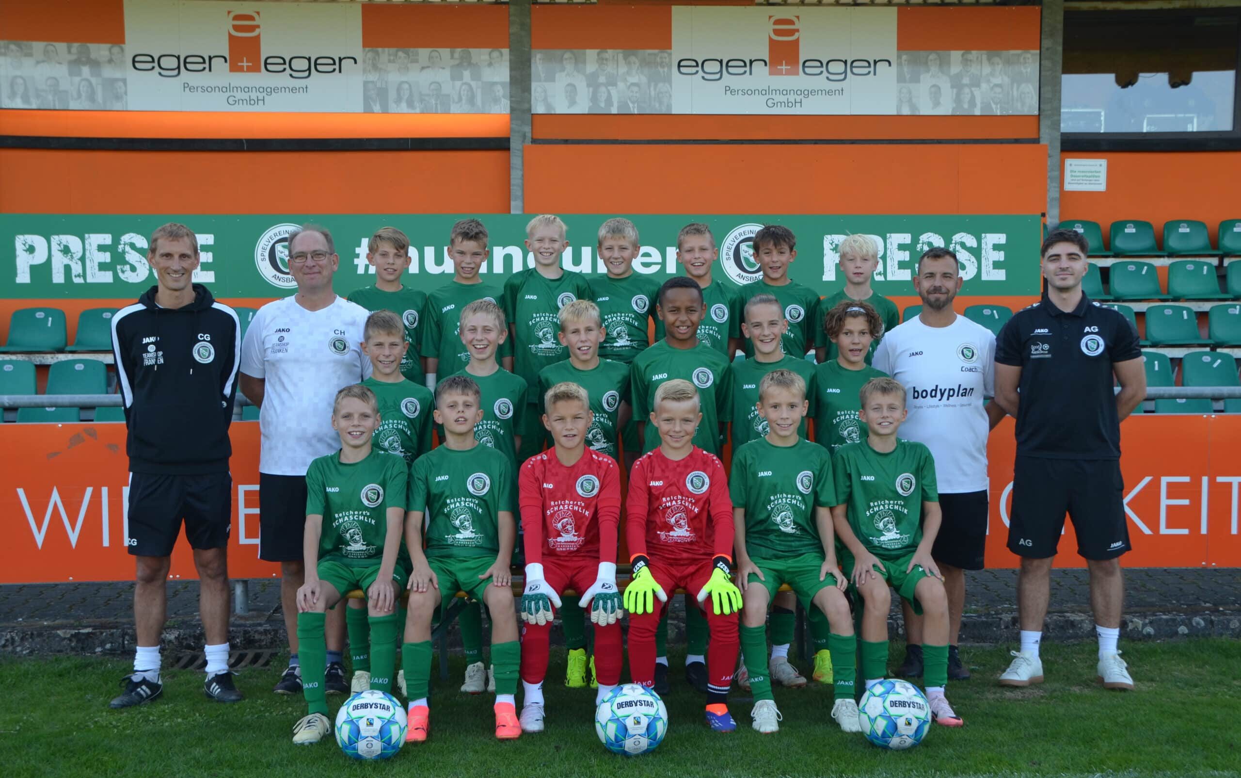 Eine Jugendfußballmannschaft posiert für ein Gruppenfoto auf einem Feld. Die Mannschaftsmitglieder in grünen Trikots stehen neben ihren Trainern. Zwei Jungen in roten Torwart-Outfits sitzen mit Fußbällen davor. Im Hintergrund sind Stadionsitze und Werbung zu sehen.